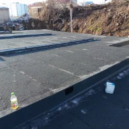 Płaski dach pokryty papą termozgrzewalną z widocznymi obróbkami blacharskimi, butelka soku i pojemnik z płynem na powierzchni, w tle budynki i skarpa.