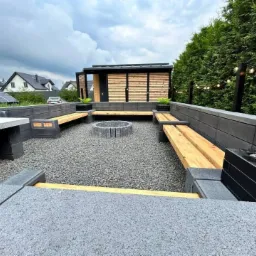 Nowoczesna aranżacja przestrzeni ogrodowej z betonowych elementów: siedziska z drewnianymi deskami, palenisko otoczone gresem, minimalistyczna altana z drewnianą elewacją i dekoracyjne oświetlenie...