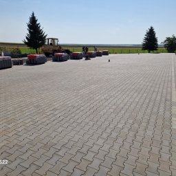 BRUKMAR MARCIN PTAK - Ukończona nawierzchnia z szarej kostki brukowej ułożonej we wzór jodełki, z przygotowanymi paletami kostki w tle i pracującą koparką oraz ekipą robotników. Widok na rozległe pole i błękitne niebo.