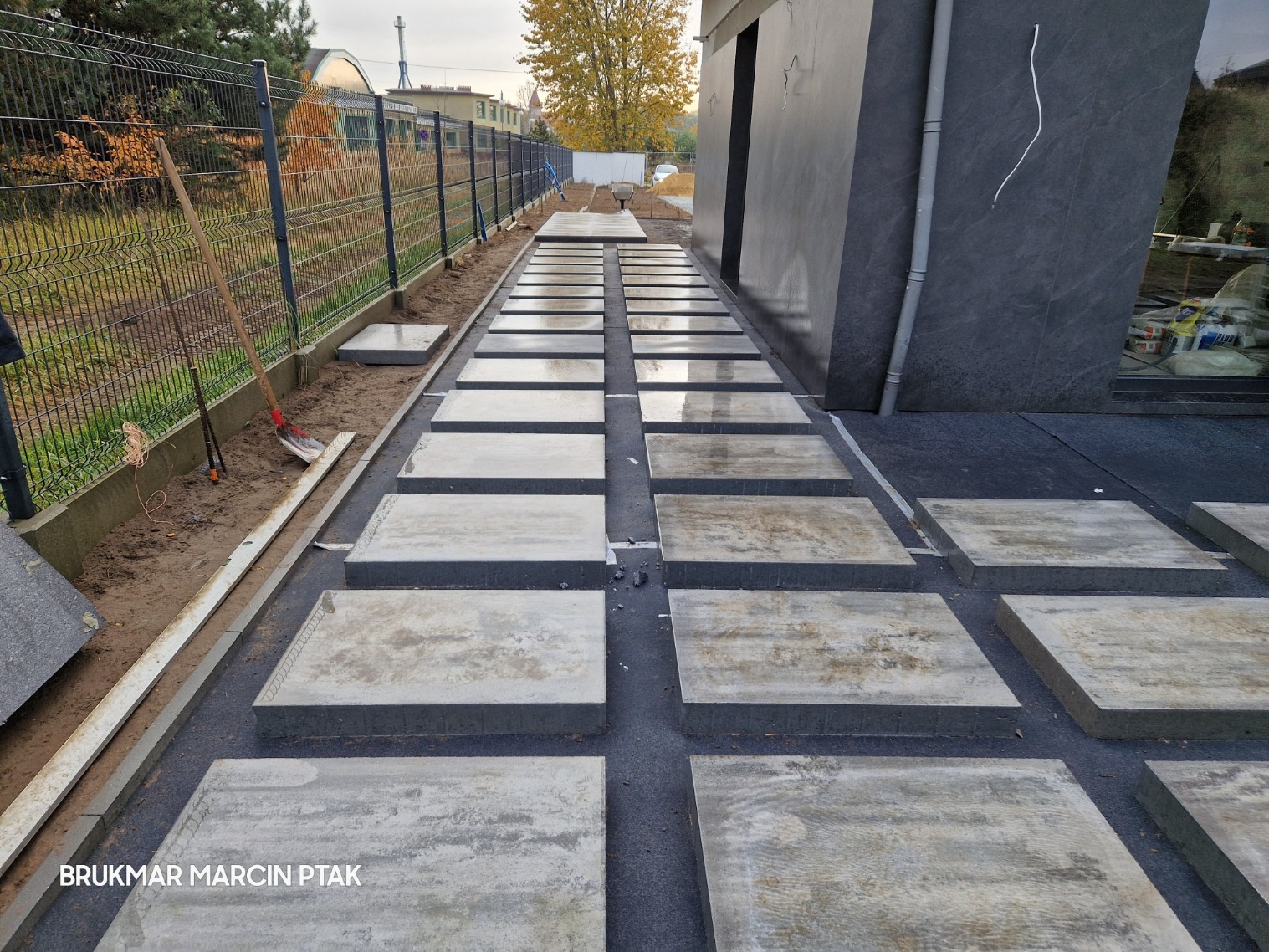 Świeżo ułożone, kwadratowe płyty chodnikowe z betonu architektonicznego tworzą ścieżkę wzdłuż nowoczesnego budynku. Widoczne narzędzia brukarskie, grunt przygotowany pod obrzeża i metalowe...