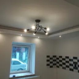 Nowoczesny pokój po remoncie w Wałbrzychu z dekoracyjną szachownicą z płytek na ścianie, widoczne oświetlenie sufitowe i okno z parapetem.