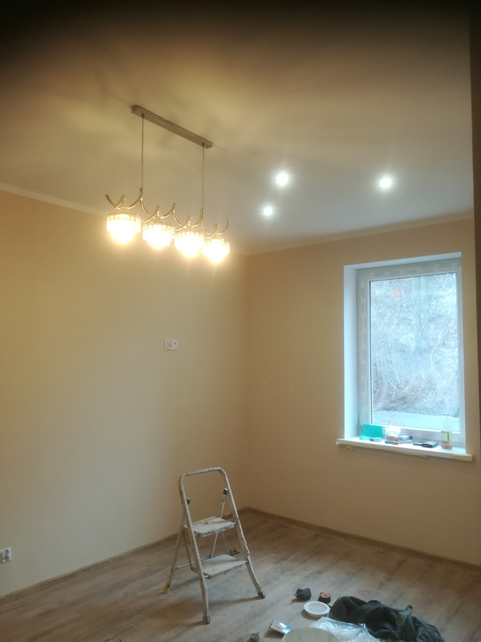 Pomalowane na beżowo wnętrze pokoju z drewnianą podłogą, widoczne okno, drabina malarska, nowoczesny żyrandol i rozproszone światło halogenowe w suficie.