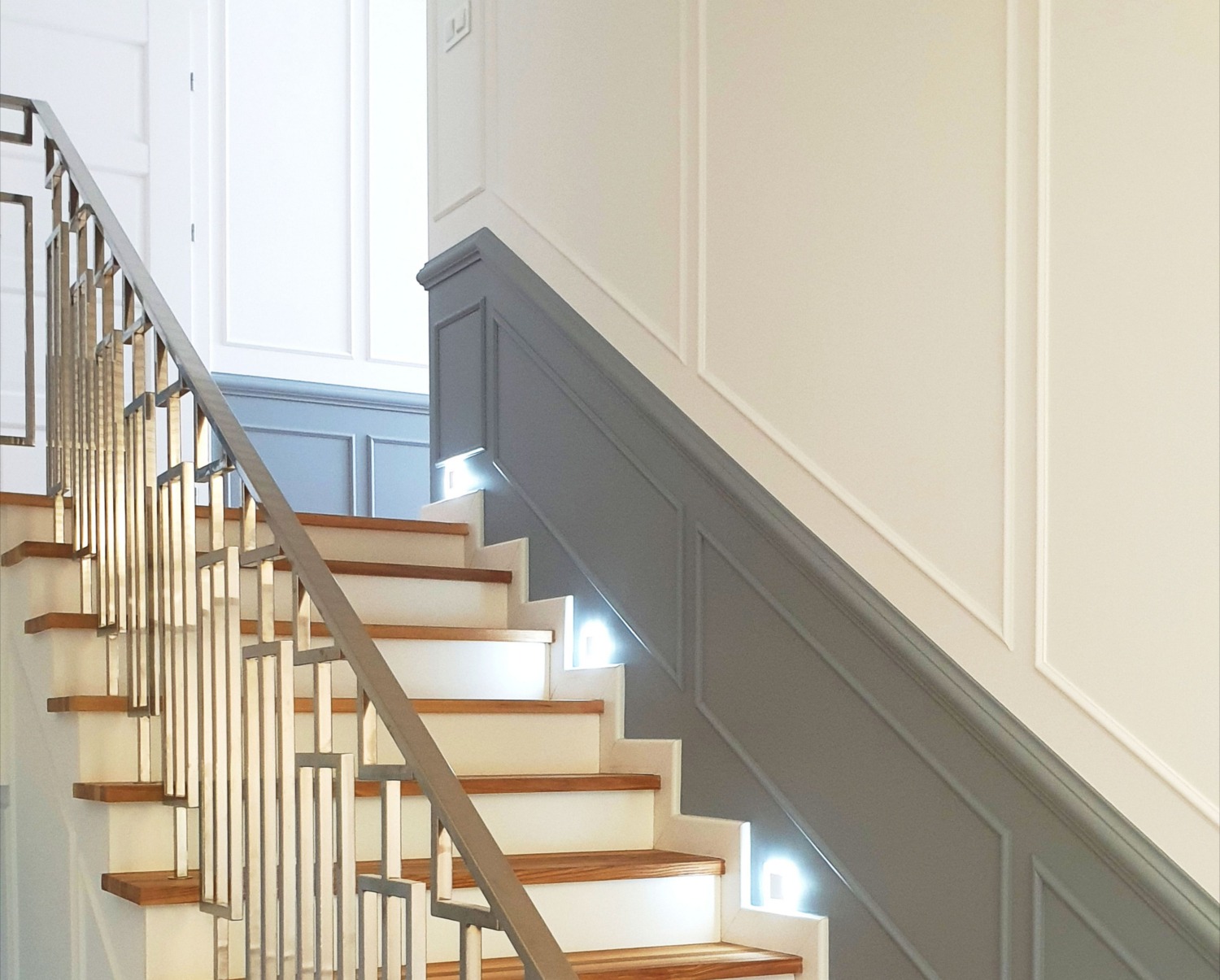 Schody z drewnianymi stopniami, białymi podstopnicami, szarą boazerią i metalową balustradą o geometrycznym wzorze, oświetlone od dołu.