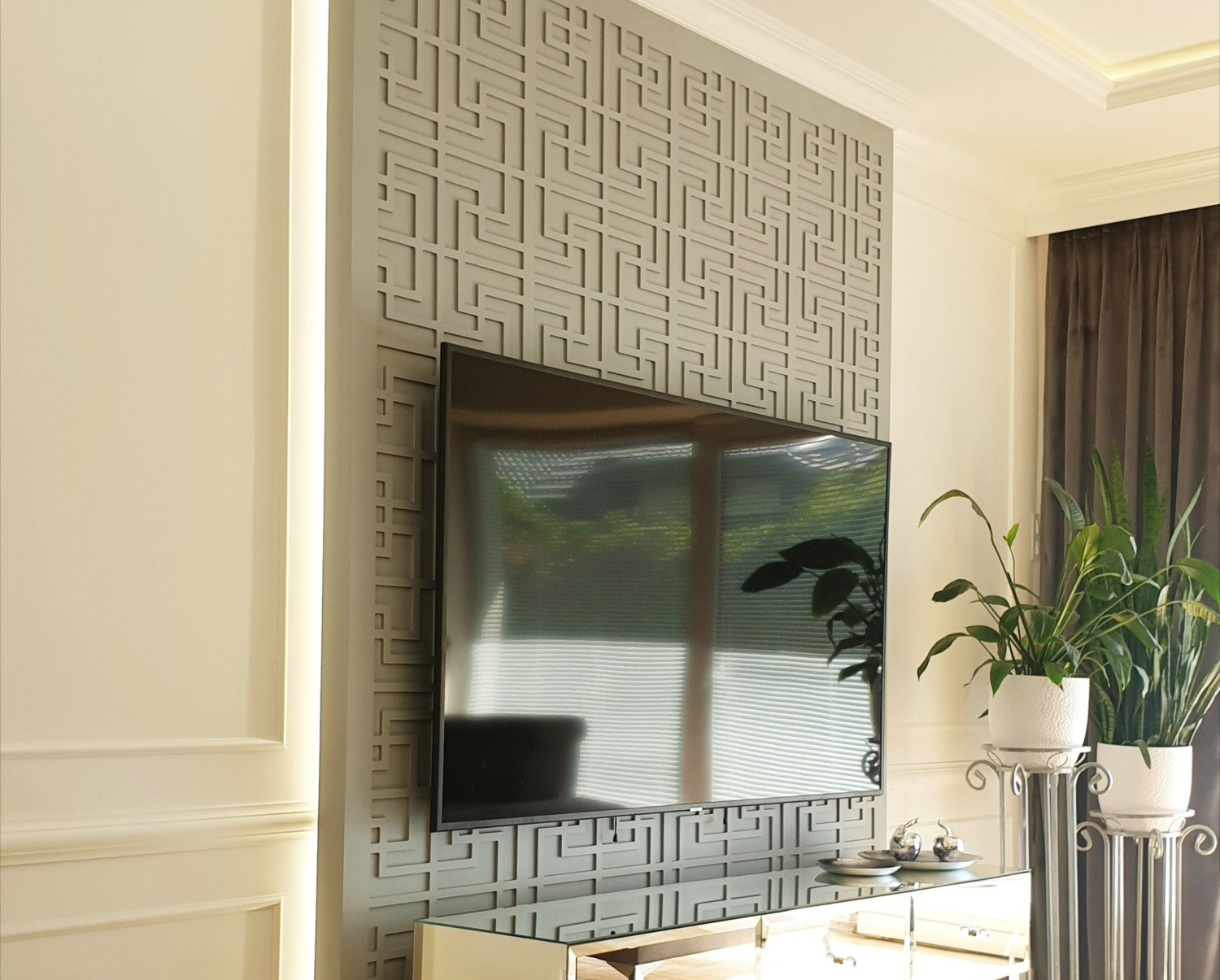 Szara ściana z dekoracyjnymi panelami 3D w geometryczny wzór, za telewizorem, w nowoczesnym salonie z roślinami doniczkowymi.