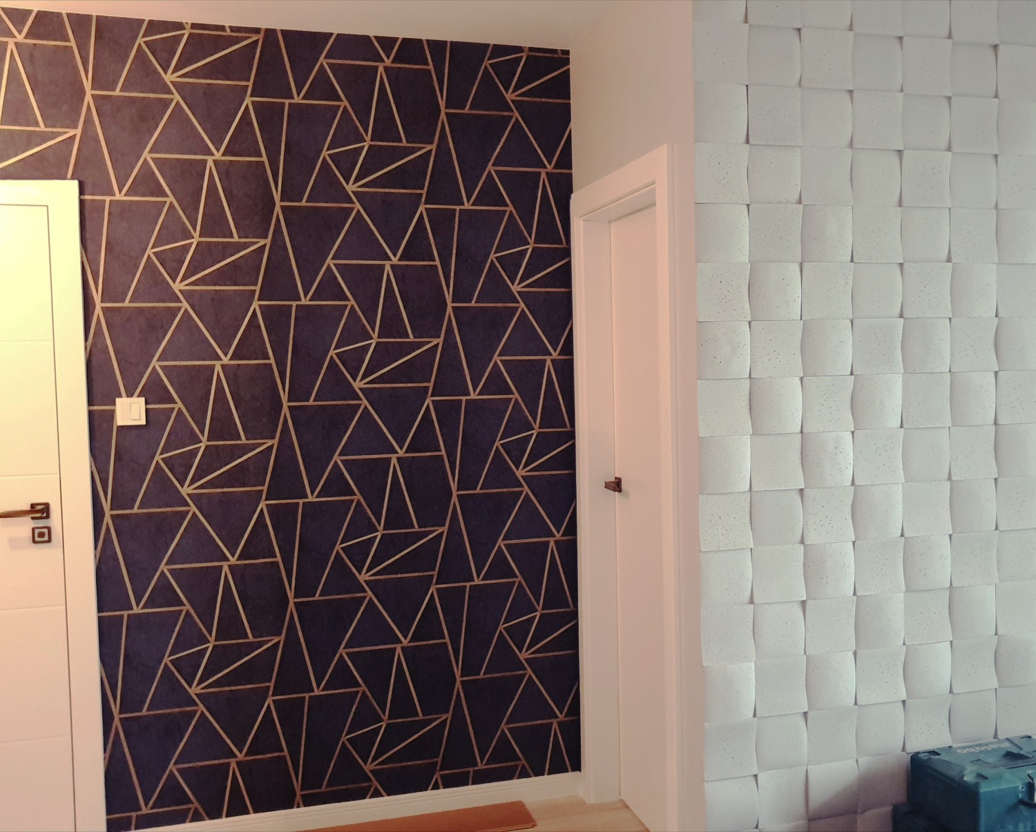 Ściana z geometrycznym wzorem w odcieniach granatu i złota sąsiadująca z białą ścianą pokrytą trójwymiarowymi panelami w kształcie kostek.