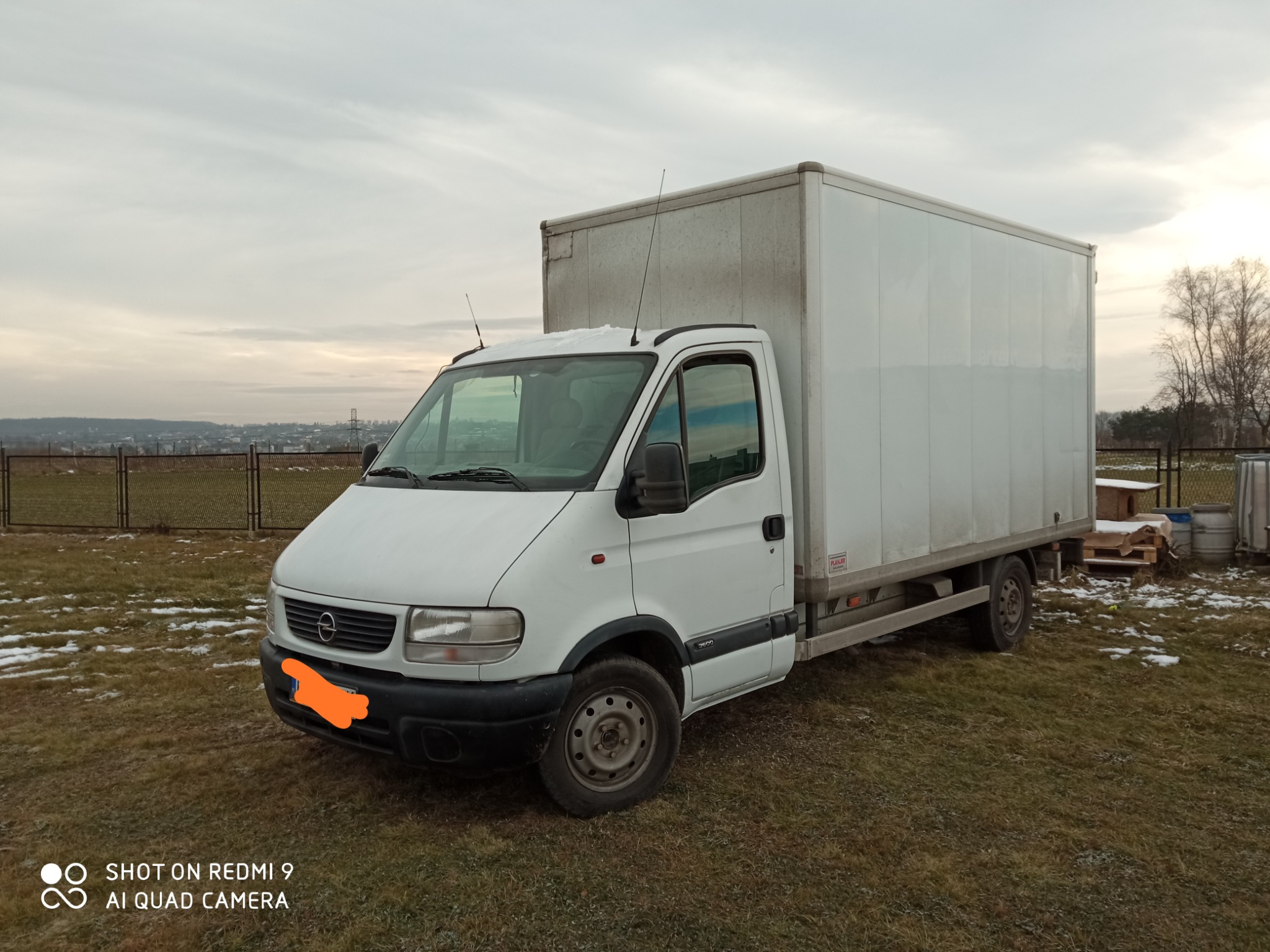 Biały samochód dostawczy z zabudową kontenerową na tle pola z resztkami śniegu i widokiem na miasto w oddali.