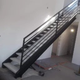 Nowoczesne schody metalowe z czarną ramą i białymi stopniami w jasnym wnętrzu, widok z dołu na górę, z widoczną osobą na górnym poziomie i elementami wykończeniowymi na podłodze.