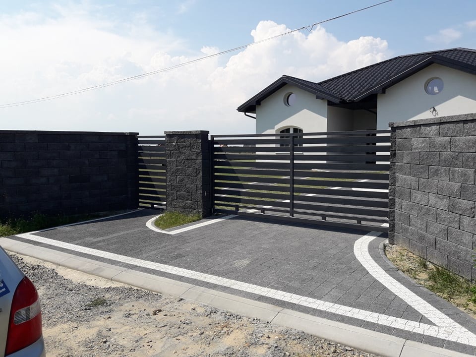 Nowoczesna posesja z przesuwną bramą wjazdową, ogrodzeniem z paneli poziomych i kostką brukową w odcieniach szarości z białymi akcentami, na tle domu z ciemnym dachem.