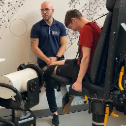 Pacjent na specjalistycznym urządzeniu do rehabilitacji kolana, nadzorowany przez rehabilitanta. Nowoczesne wyposażenie gabinetu, logo firmy widoczne na koszulce.