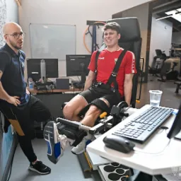 Pacjent uśmiechnięty, w czerwonej koszulce, ćwiczy nogę na specjalistycznej maszynie pod nadzorem fizjoterapeuty w nowoczesnym gabinecie rehabilitacyjnym.