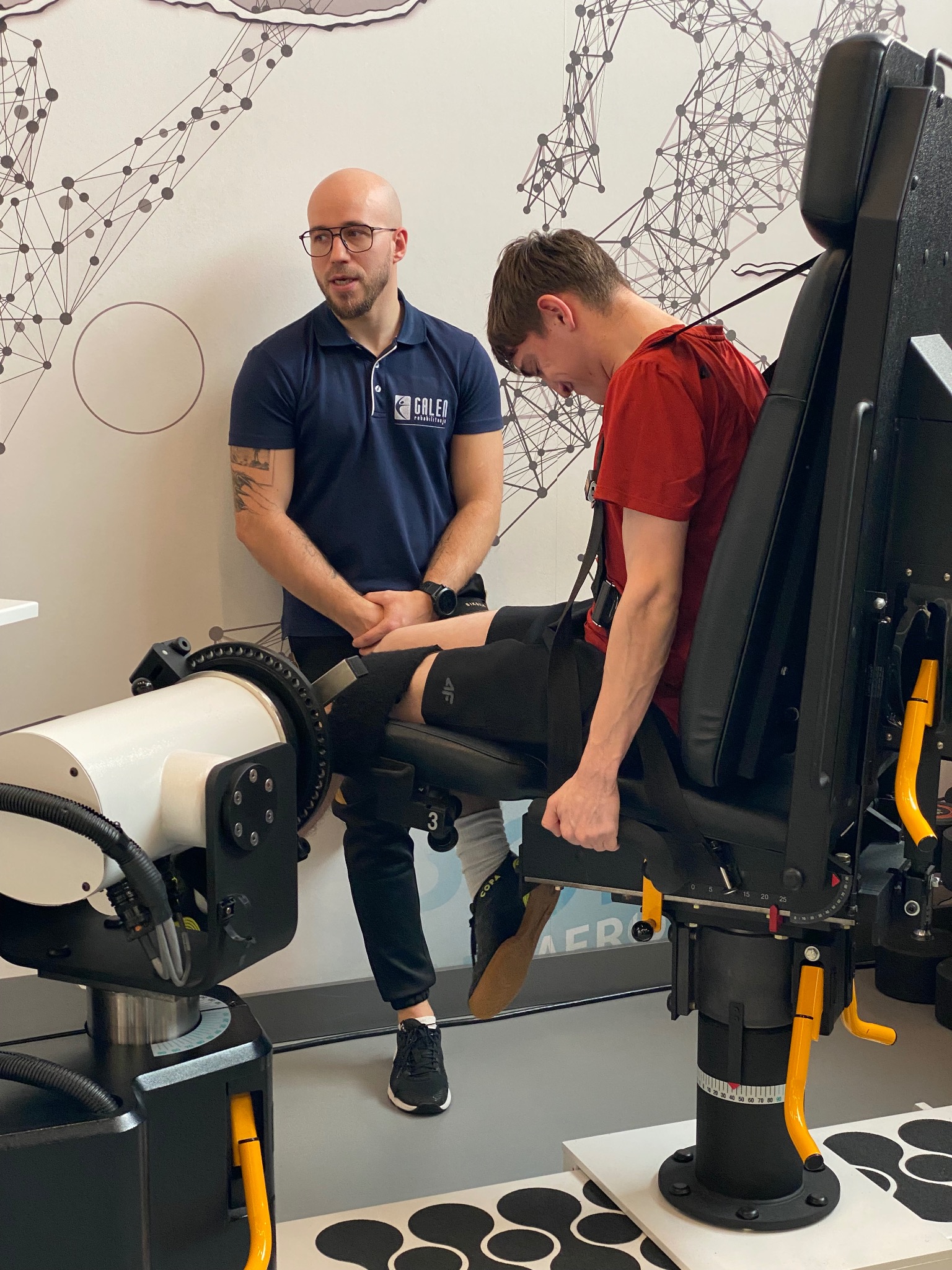 Pacjent na specjalistycznym urządzeniu do rehabilitacji kolana, nadzorowany przez rehabilitanta. Nowoczesne wyposażenie gabinetu, logo firmy widoczne na koszulce.
