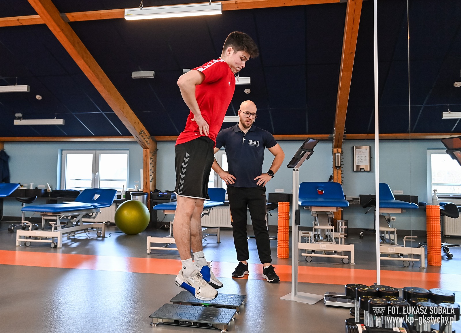 Zawodnik w trakcie testu skoczności na platformie dynamometrycznej, nadzorowany przez specjalistę w gabinecie z leżankami i sprzętem do ćwiczeń.