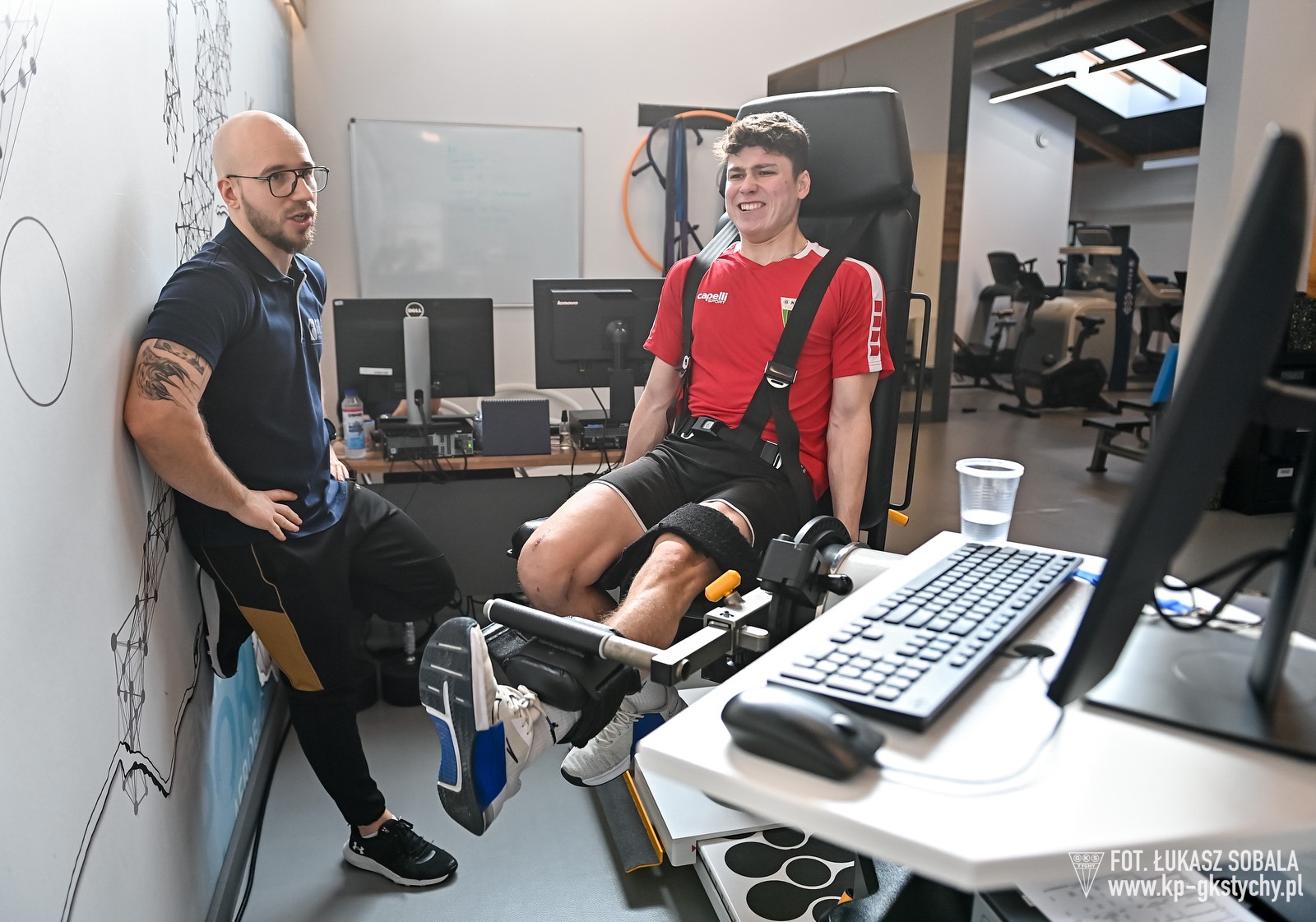 Pacjent uśmiechnięty, w czerwonej koszulce, ćwiczy nogę na specjalistycznej maszynie pod nadzorem fizjoterapeuty w nowoczesnym gabinecie rehabilitacyjnym.