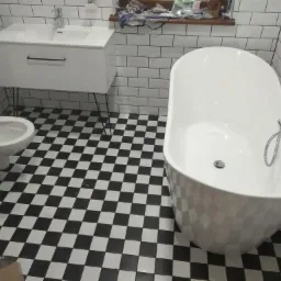 Nowoczesna łazienka z białą wanną wolnostojącą, umywalką na metalowych nóżkach i toaletą na tle czarno-białej szachownicy na podłodze i białych płytek na ścianach.