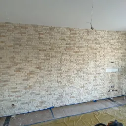 Ściana wyłożona dekoracyjną cegłą w Siekierczynie, z widocznymi puszkami elektrycznymi i przygotowanym miejscem pod telewizor, zabezpieczona folią podłoga i drabina w tle.