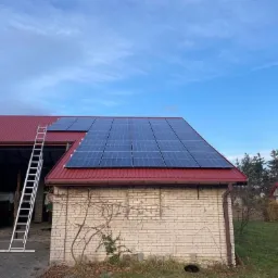 Panele słoneczne na czerwonym dachu budynku gospodarczego z otwartymi wrotami, drabina oparta o ścianę, błękitne niebo w tle.