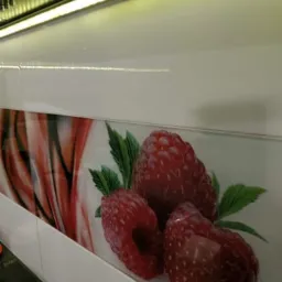 Białe płytki ścienne w kuchni z dekoracyjnym motywem malin i abstrakcyjnym wzorem, oświetlone od góry listwą LED.