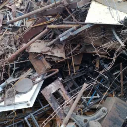 Stos pordzewiałych metalowych odpadów i złomu, w tym rury, blachy, elementy konstrukcyjne, z widocznymi białymi beczkami w tle po lewej stronie i logo firmy w prawym dolnym rogu.