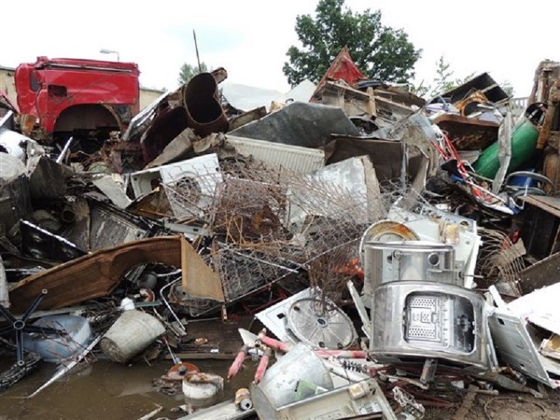Sterta złomu i odpadów budowlanych, w tym fragmenty karoserii czerwonej ciężarówki, metalowe elementy pralek, grzejniki i inne materiały, zalegające na błotnistym terenie.