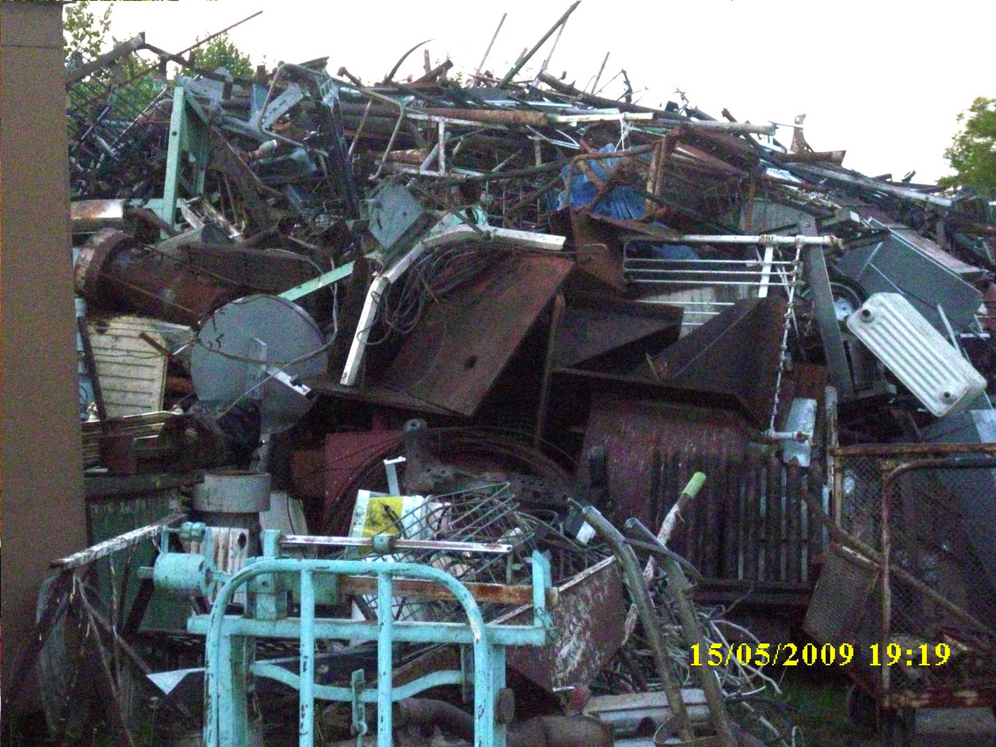 Sterta pordzewiałych metalowych odpadów, w tym rury, grzejniki, elementy konstrukcyjne i metalowe kraty, sfotografowana w słabym oświetleniu z widoczną datą 15/05/2009.