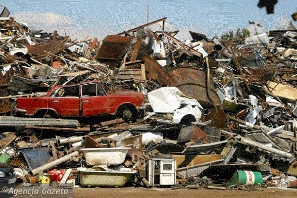 Góra złomu z widocznym czerwonym samochodem osobowym, zgniecionym białym autem, fragmentami wanien, blach i innych metalowych odpadów pod błękitnym niebem.