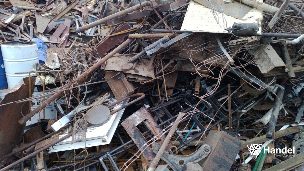 Stos pordzewiałych metalowych odpadów i złomu, w tym rury, blachy, elementy konstrukcyjne, z widocznymi białymi beczkami w tle po lewej stronie i logo firmy w prawym dolnym rogu.