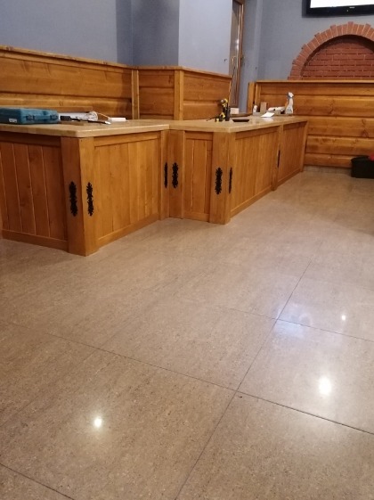 Wykończone drewniane panele ścienne i zabudowa baru z dekoracyjnymi, kutymi okuciami w lokalu z jasną, kamienną posadzką. Częściowo widoczna ściana z ceglanym łukiem.