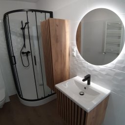 Jacek Morawski Usługi Remontowo-Budowlane ALANIS - Nowoczesna łazienka po remoncie z kabiną prysznicową z czarnymi ramami, szafką z umywalką i lustrem z podświetleniem LED oraz drewnianymi akcentami na szafkach i podłodze.