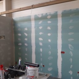 Jacek Morawski Usługi Remontowo-Budowlane ALANIS - Wnętrze w trakcie wykańczania: płyty karton gips z widocznymi śladami szpachlowania i wstawionymi puszkami elektrycznymi, narzędzia i materiały budowlane na pierwszym planie, drewniana konstrukcja...