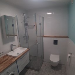 Jacek Morawski Usługi Remontowo-Budowlane ALANIS - Nowoczesna łazienka po remoncie z kabiną prysznicową, umywalką nablatową na drewnianym blacie, podwieszaną toaletą i pralką pod blatem, wykończona białymi płytkami i wzorzystą podłogą.