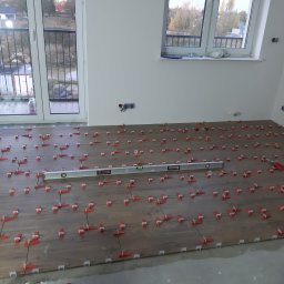 Jacek Morawski Usługi Remontowo-Budowlane ALANIS - Układanie płytek imitujących drewno w jasnym pomieszczeniu, widoczny poziomica i czerwone kliny dystansowe, okno balkonowe z widokiem na zewnątrz.