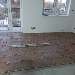 Układanie płytek podłogowych imitujących drewno, z użyciem czerwonych klinów poziomujących i poziomicy; widoczne gniazdka elektryczne na ścianie i okno balkonowe.