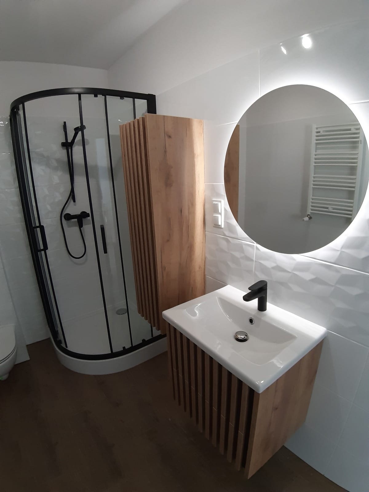 Nowoczesna łazienka po remoncie z kabiną prysznicową z czarnymi ramami, szafką z umywalką i lustrem z podświetleniem LED oraz drewnianymi akcentami na szafkach i podłodze.