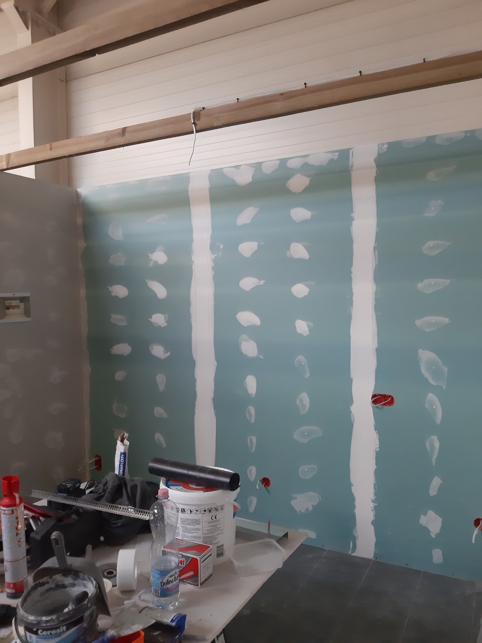 Wnętrze w trakcie wykańczania: płyty karton gips z widocznymi śladami szpachlowania i wstawionymi puszkami elektrycznymi, narzędzia i materiały budowlane na pierwszym planie, drewniana konstrukcja...