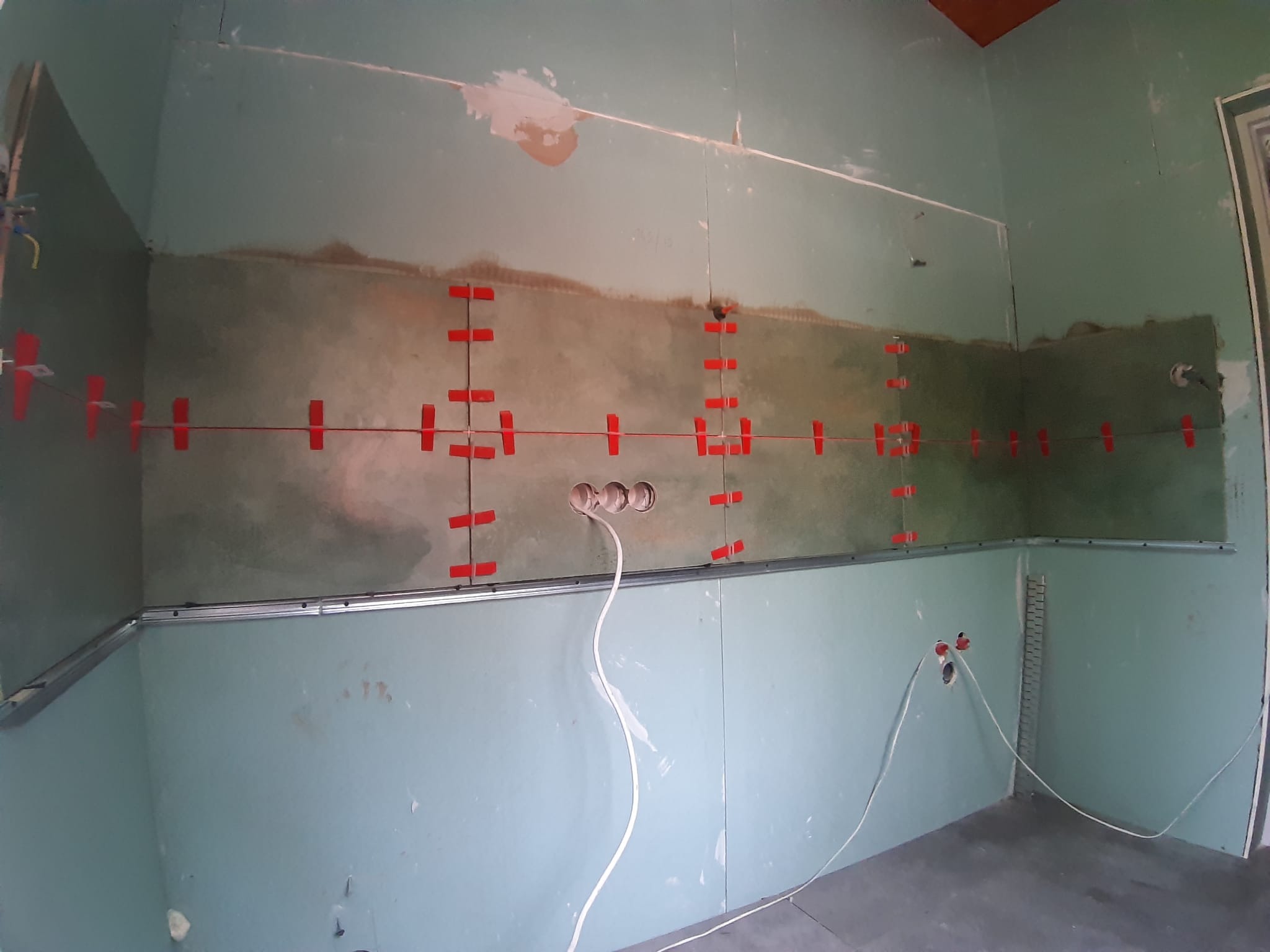 Przygotowanie ściany z płyty karton gips do układania płytek, widoczne profile metalowe, wyprowadzone przewody elektryczne i czerwone krzyżyki do poziomowania płytek.