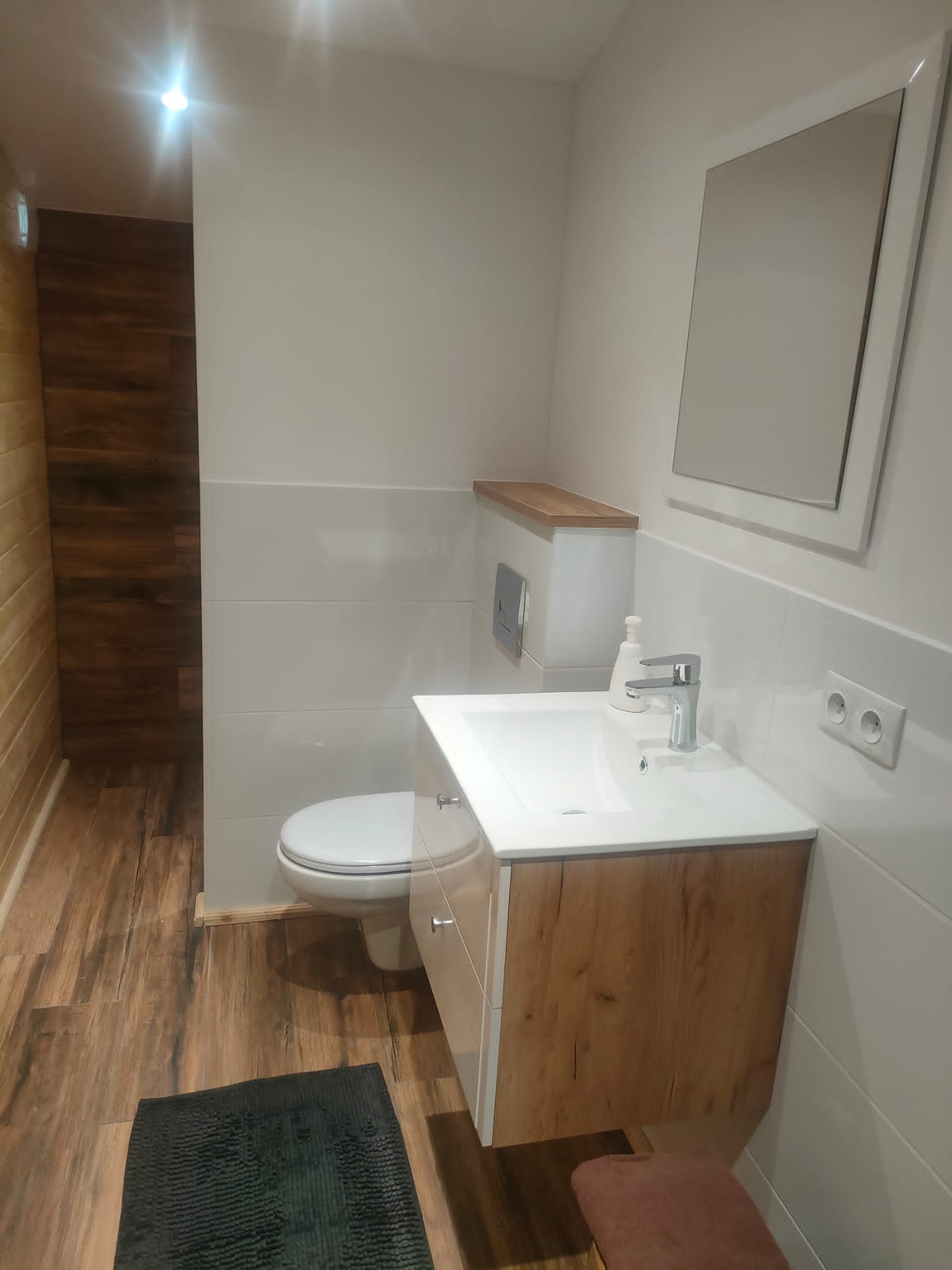 Wykończona łazienka z drewnopodobną podłogą i szafką pod umywalką, białe płytki ścienne do połowy wysokości, toaleta kompaktowa i prostokątne lustro nad umywalką.