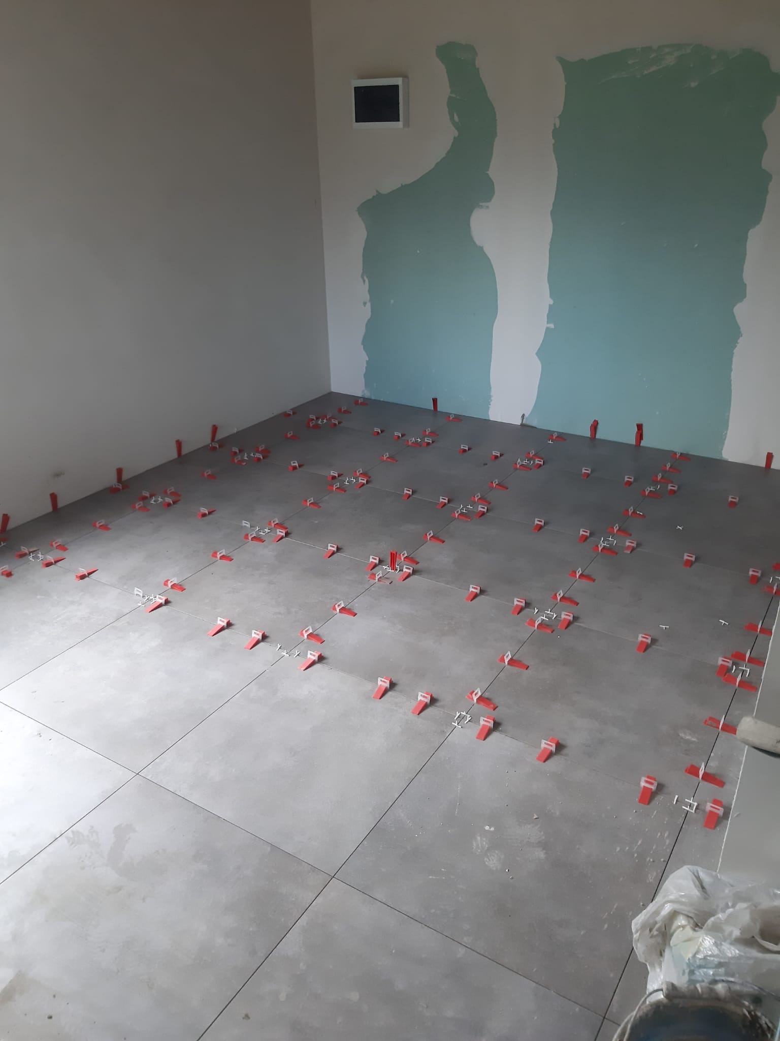 Szare płytki podłogowe w trakcie układania, z użyciem czerwonych klinów poziomujących, na ścianie widoczne ślady po szpachlowaniu.