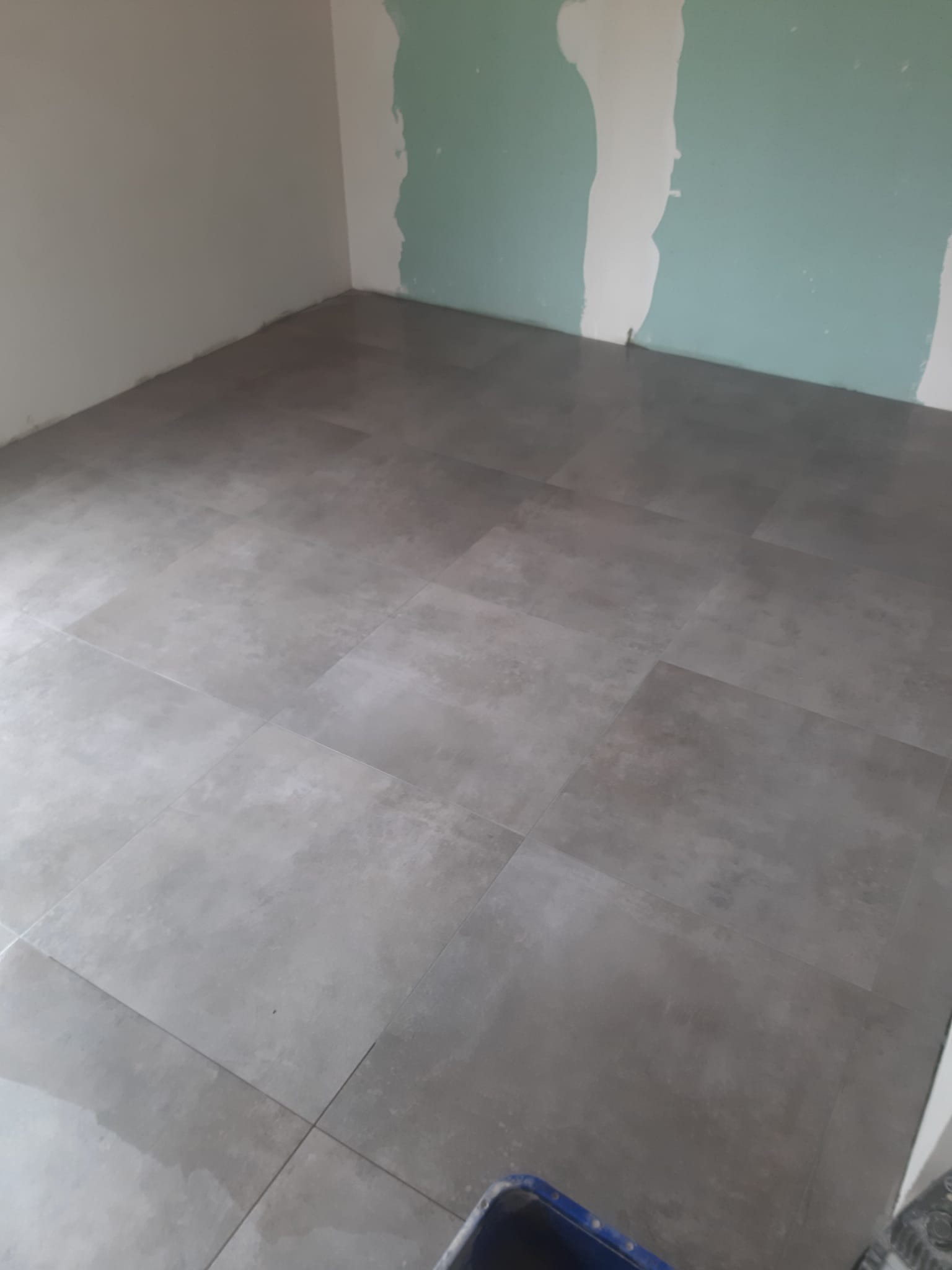 Ułożone szare płytki podłogowe w nowo wykończonym pomieszczeniu z fragmentami ścian przygotowanych pod malowanie i widocznym narożnikiem niebieskiego wiadra.