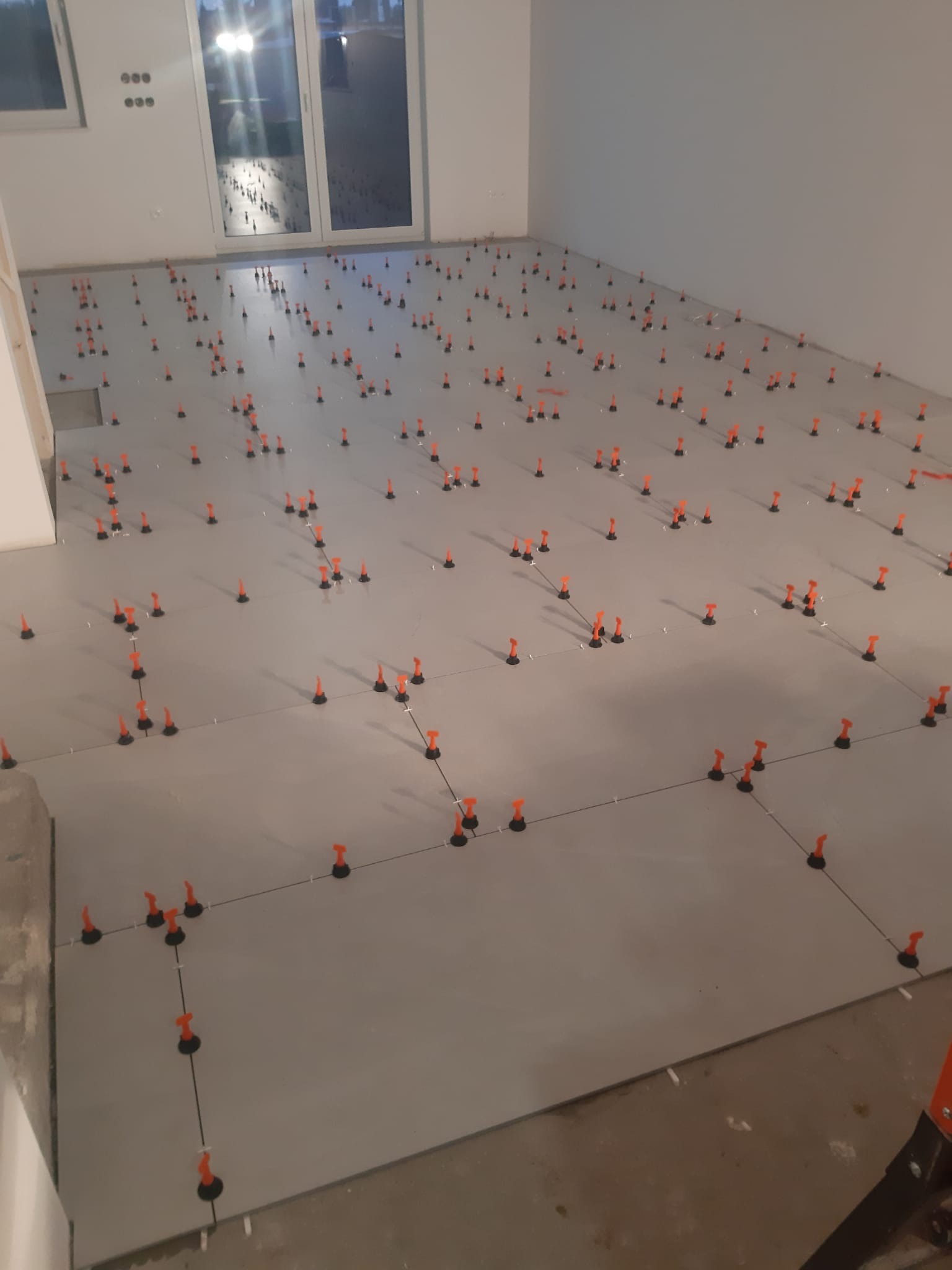 Układanie jasnoszarych płytek podłogowych z użyciem pomarańczowych klipsów poziomujących, widok z góry na niedokończoną podłogę w jasnym pomieszczeniu.