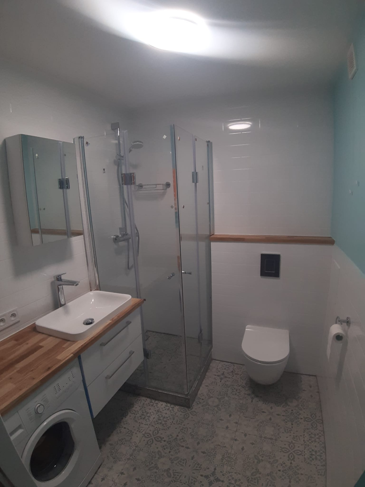 Nowoczesna łazienka po remoncie z kabiną prysznicową, umywalką nablatową na drewnianym blacie, podwieszaną toaletą i pralką pod blatem, wykończona białymi płytkami i wzorzystą podłogą.