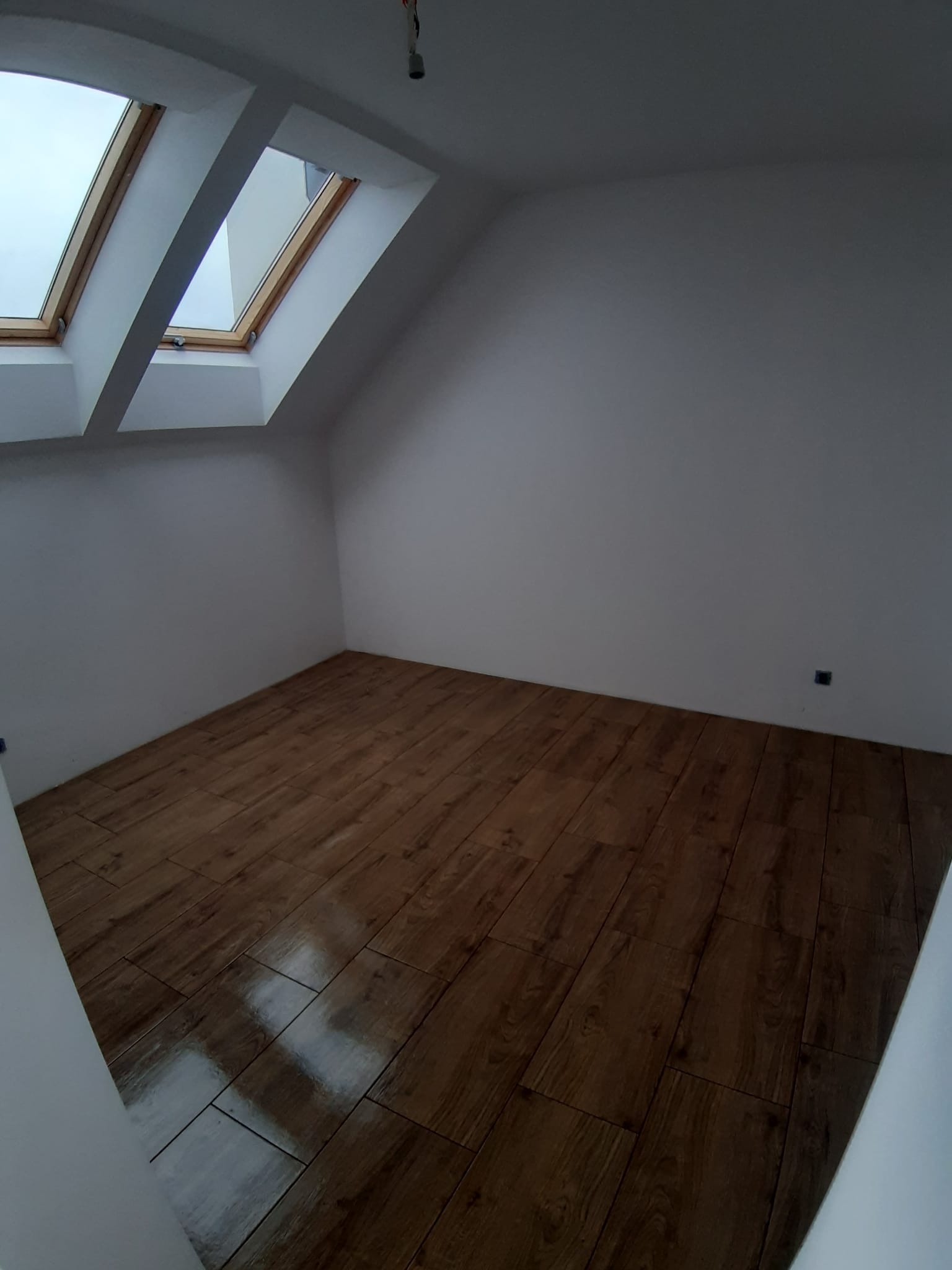 Poddasze w Poznaniu po remoncie, z brązowymi płytkami imitującymi drewno na podłodze i dwoma oknami dachowymi w skośnym suficie.
