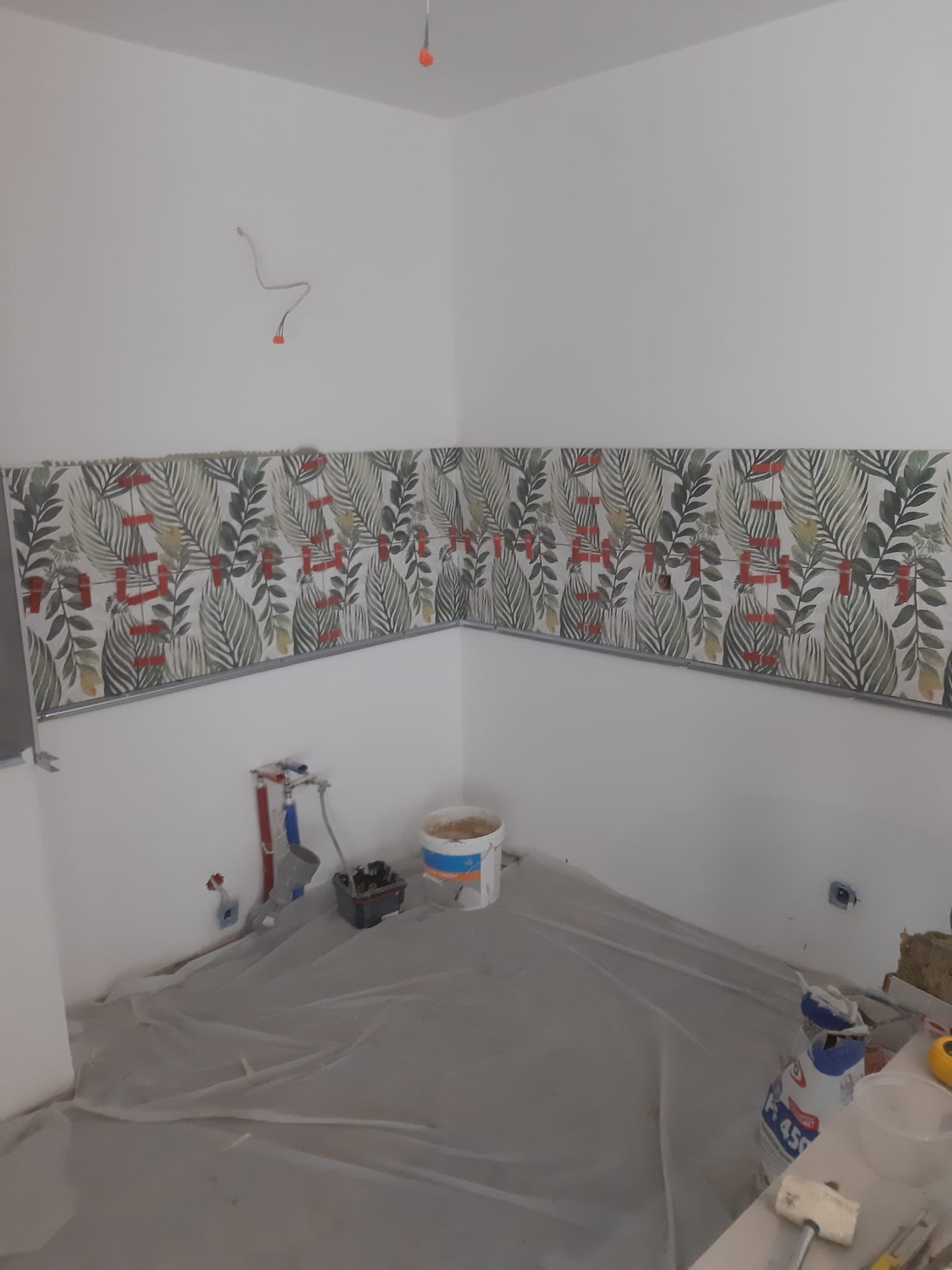 Fragment ściany w trakcie układania płytek z motywem roślinnym, widoczne narzędzia i materiały budowlane, zabezpieczona folią podłoga.