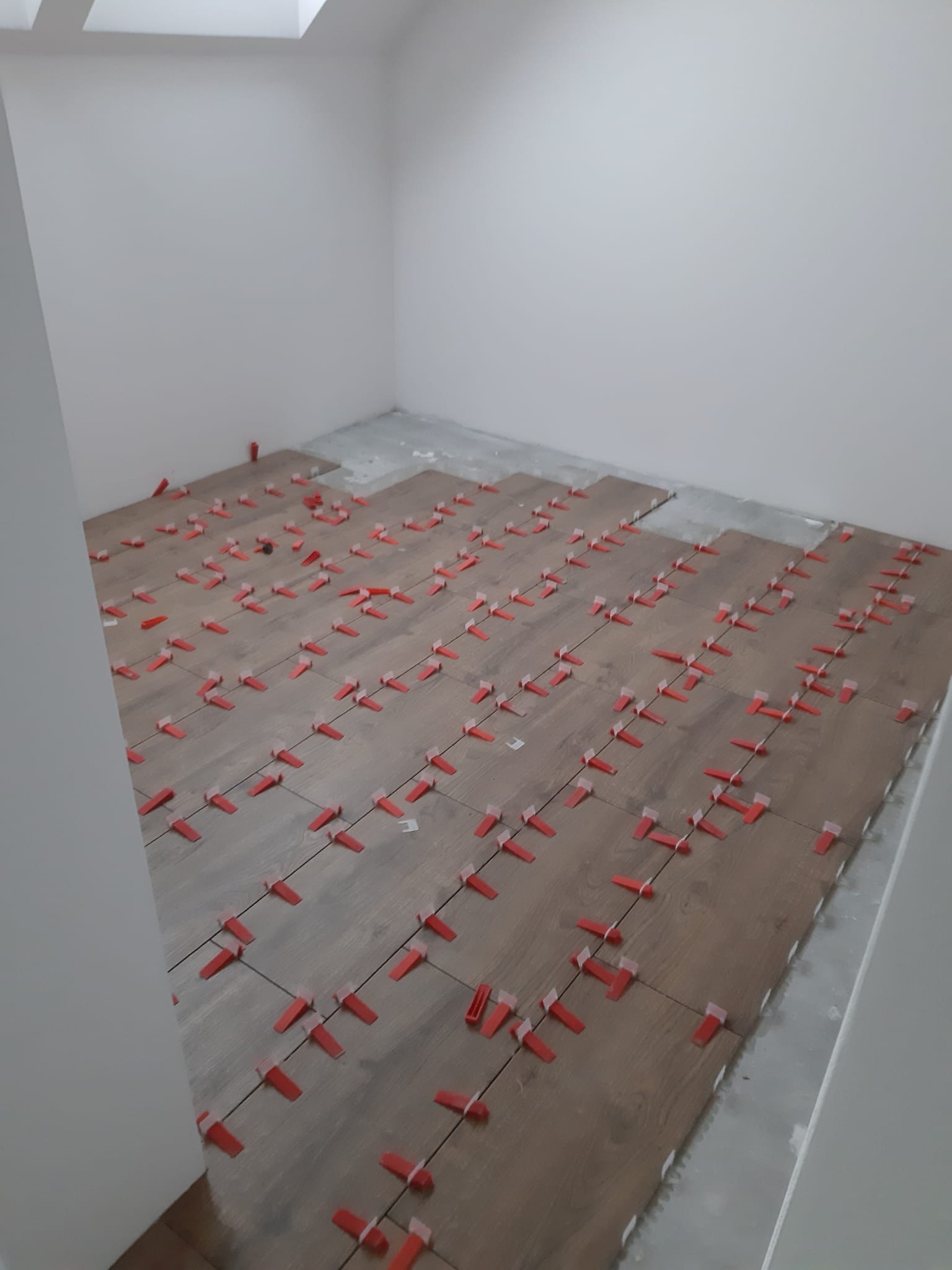 Układanie płytek podłogowych imitujących drewno w jasnym pomieszczeniu z użyciem czerwonych klinów poziomujących; widoczny fragment surowej podłogi.