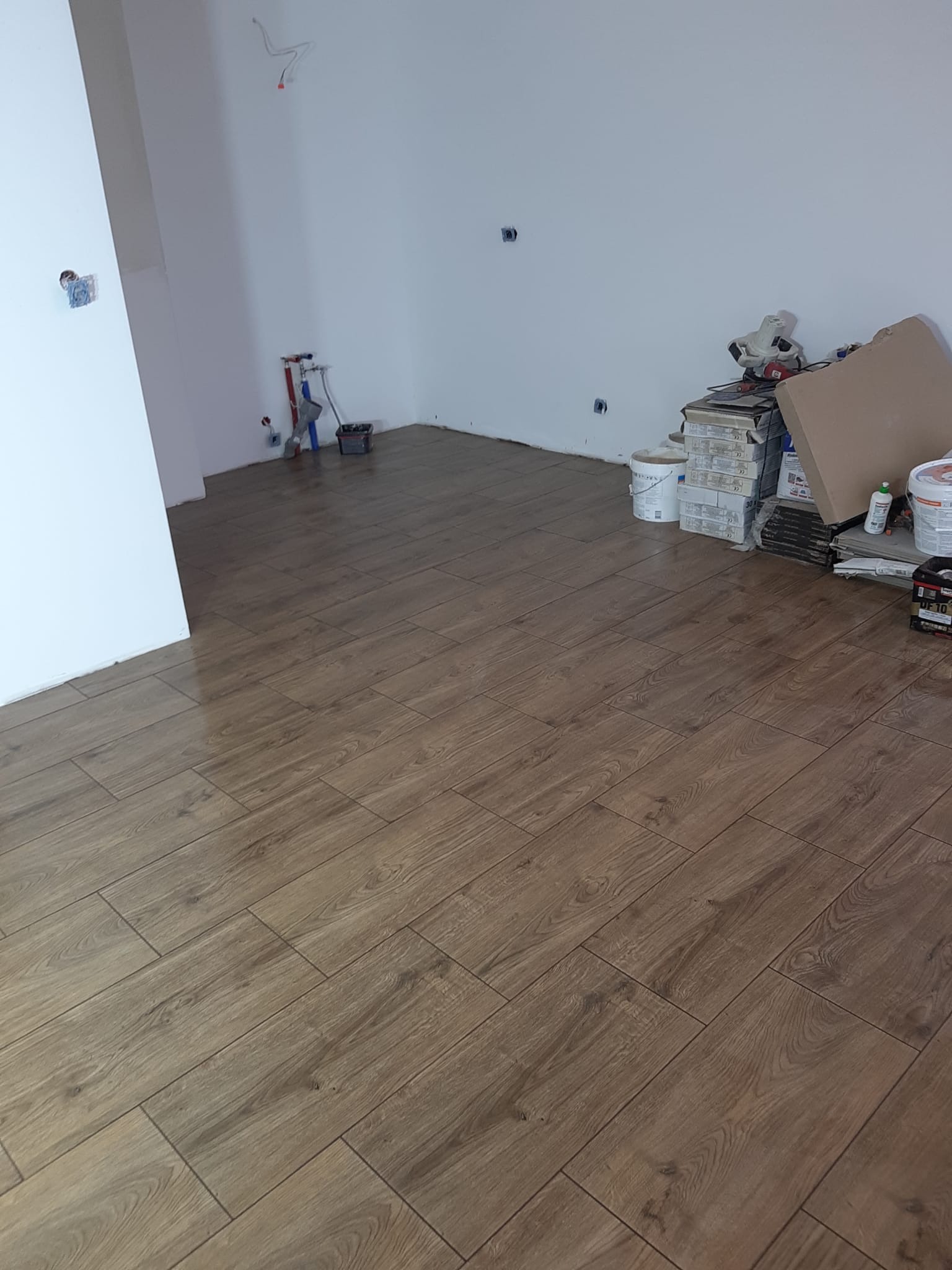 Ułożona podłoga z płytek imitujących drewno w pustym pokoju, widoczne narzędzia i materiały budowlane w tle, instalacja wodna przy ścianie.