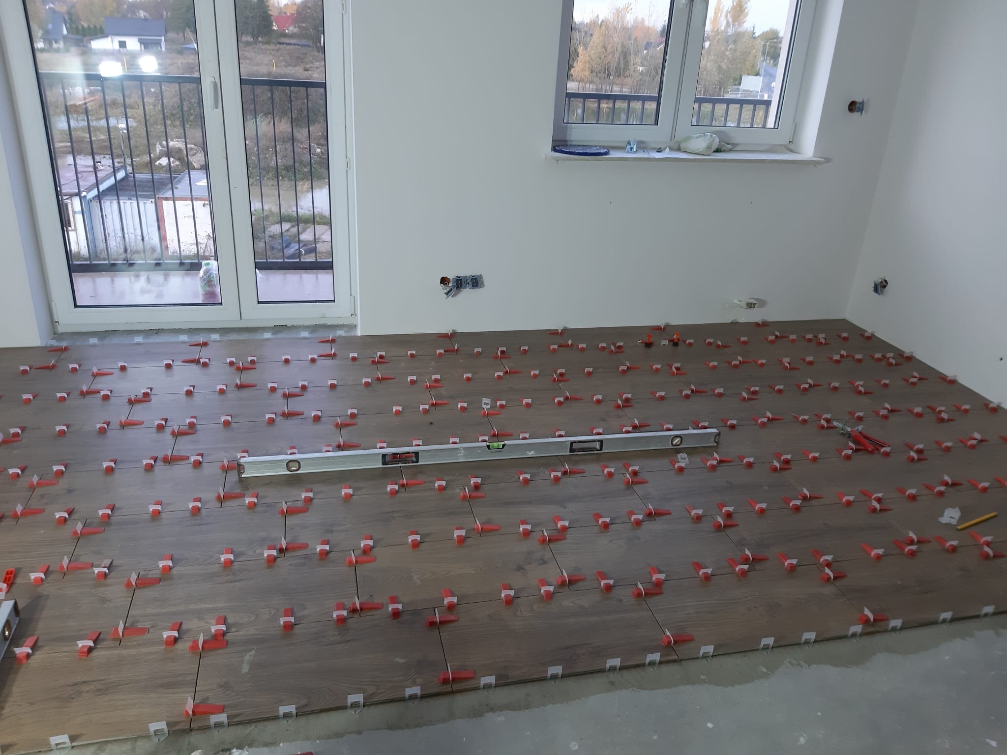 Układanie płytek podłogowych imitujących drewno, z użyciem czerwonych klinów poziomujących i poziomicy; widoczne gniazdka elektryczne na ścianie i okno balkonowe.