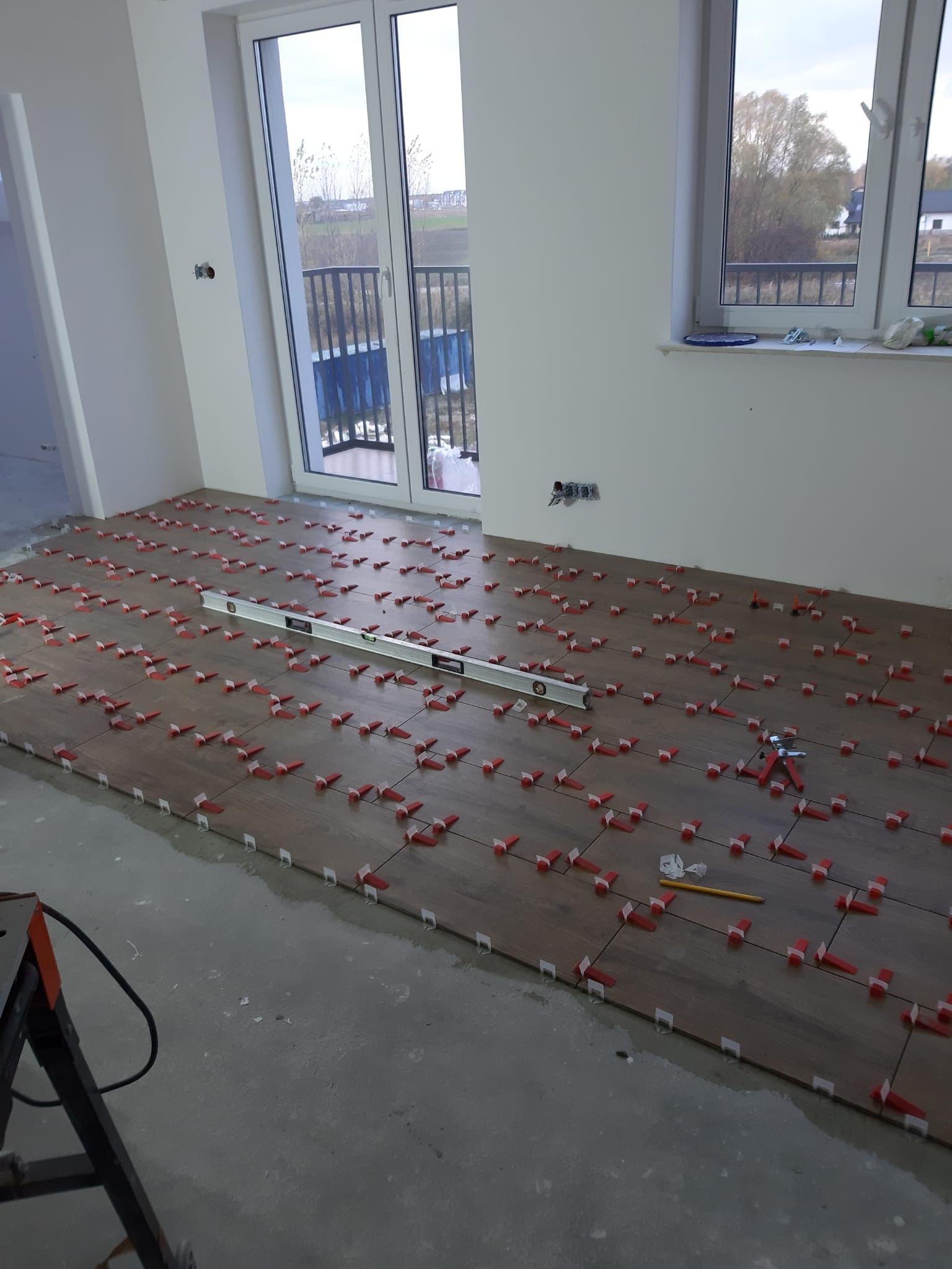 Układanie podłogi z płytek imitujących drewno, z użyciem poziomicy i czerwonych klinów dystansowych, w jasnym, nowym pomieszczeniu z dużym oknem balkonowym.