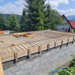 Wznoszenie fundament&oacute;w z szalunkiem i zbrojeniem na tle g&oacute;rskiego krajobrazu, widoczny betonowy murek, siatka zbrojeniowa i drewniany szalunek.