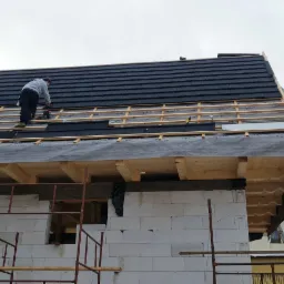 Montaż czarnej blachodachówki na drewnianej konstrukcji dachu domu z białego bloczka, widoczni dekarze przy pracy.