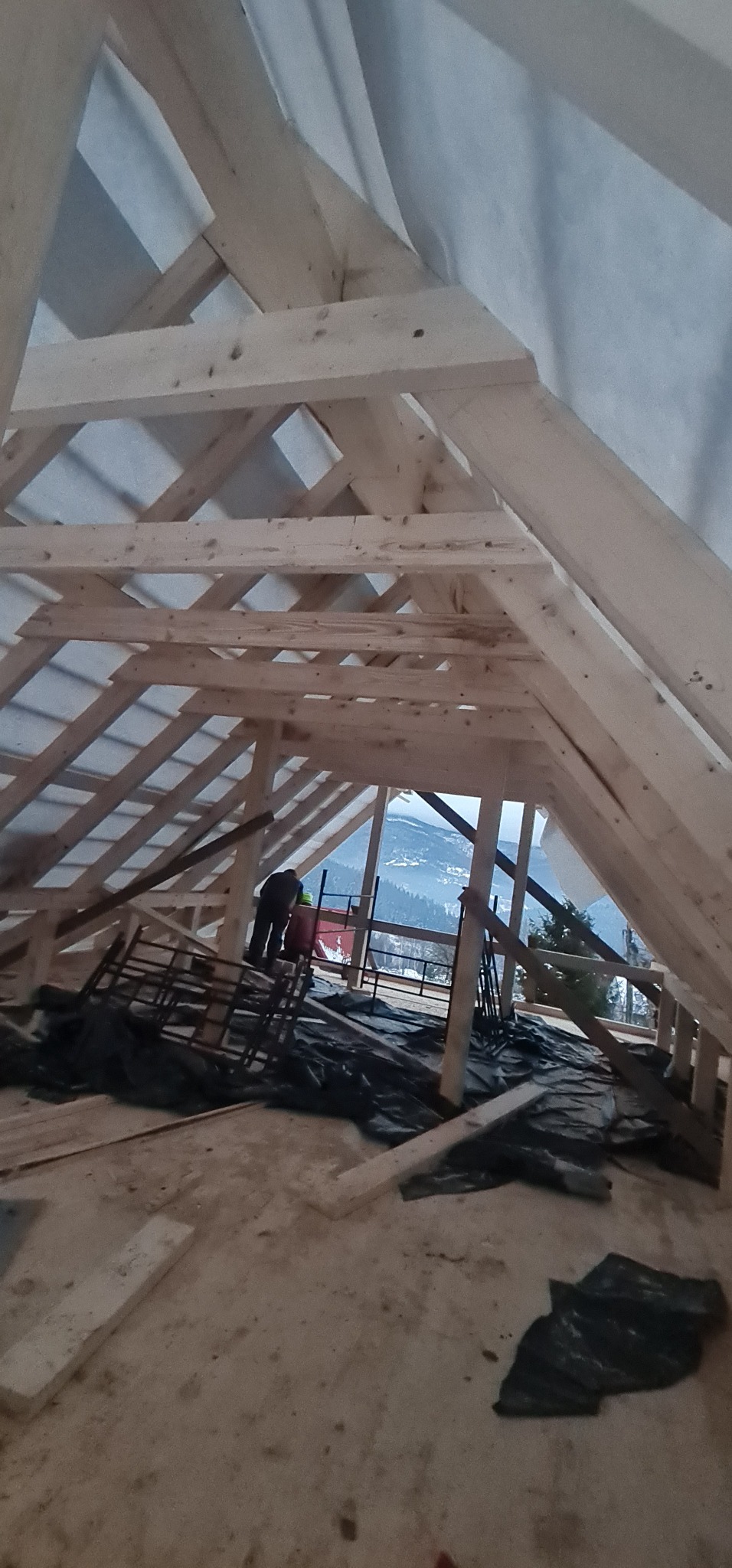 Wnętrze domu w trakcie budowy, widoczna drewniana konstrukcja dachu z folią, w tle góry i pracownicy w kaskach.