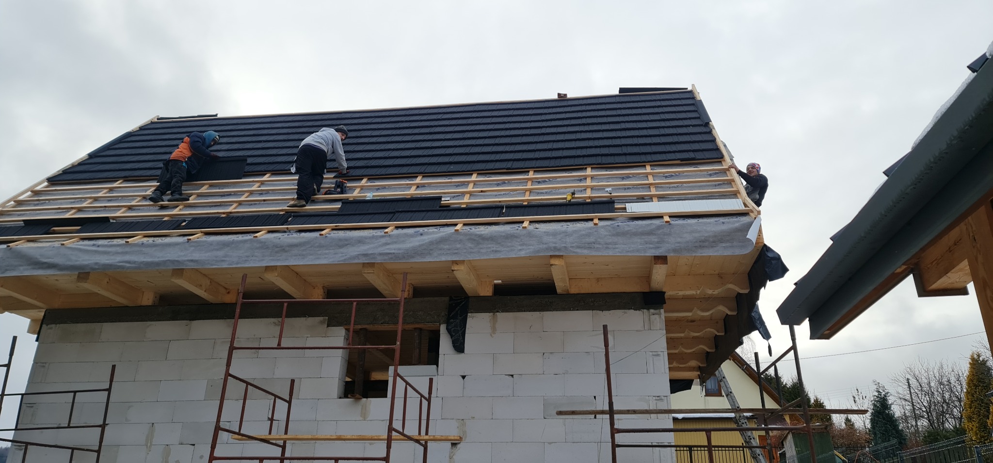 Montaż czarnej blachodachówki na drewnianej konstrukcji dachu domu z białego bloczka, widoczni dekarze przy pracy.